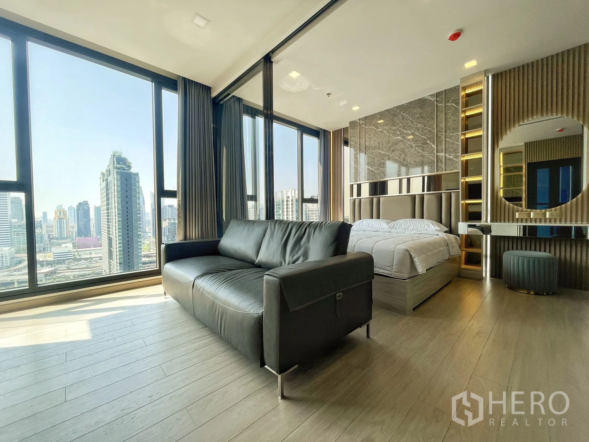 Condominium for rent in Huai Khwang, Bangkok - Bright condo with black leather sofa, bed, floor-to-ceiling windows showcasing a city skyline, built-in vanity, and wood flooring.
