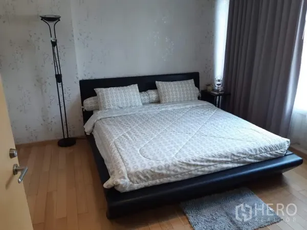 Condominium for rent in Khlong Toei, Bangkok - Cozy bedroom with a king-size platform bed, light wallpaper, bedside table and large curtained window.