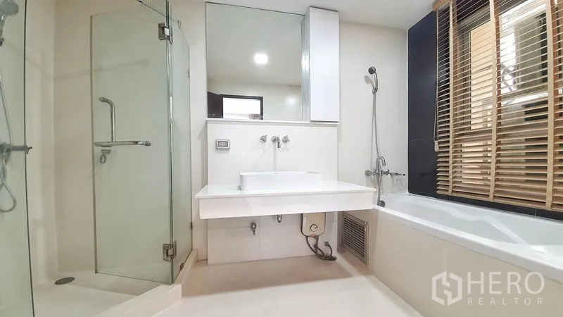 Condominium for rent in Khlong Toei, Bangkok - Primary bathroom with glass-enclosed shower, bathtub by the window with blinds, large mirror and vanity.