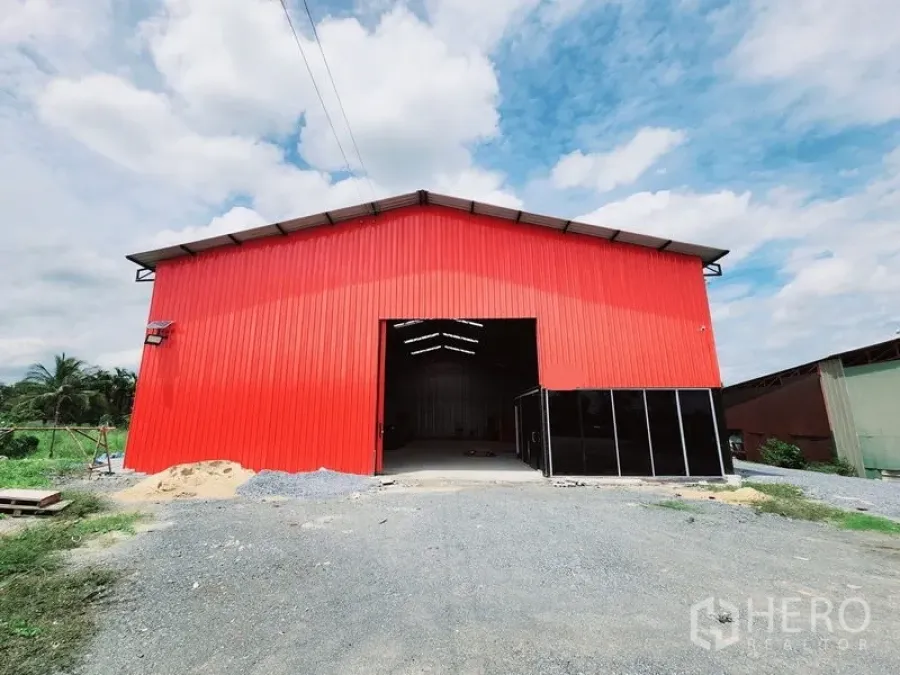 Warehouse for rent in Sam Phran, Nakhon Pathom - Approach to the red warehouse showing the open bay and gravel driveway under a cloudy sky.
