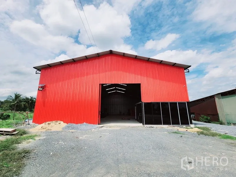 Warehouse for rent in Sam Phran, Nakhon Pathom - Approach to the red warehouse showing the open bay and gravel driveway under a cloudy sky.