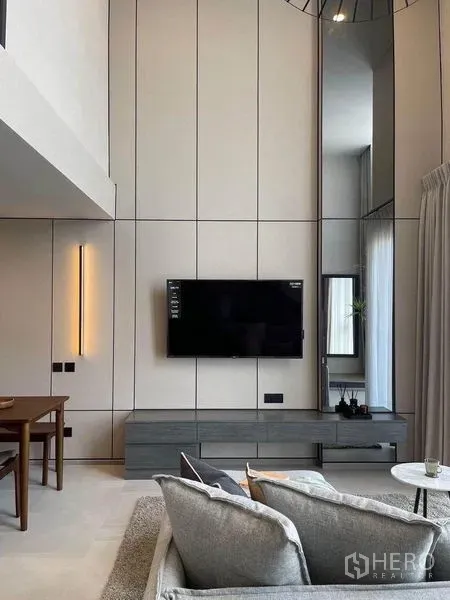 Condominium for rent in Pathum Wan, Bangkok - Modern loft living room with double-height walls, wall-mounted TV on paneling, grey sofa, media console, tall mirror and soft drapes.