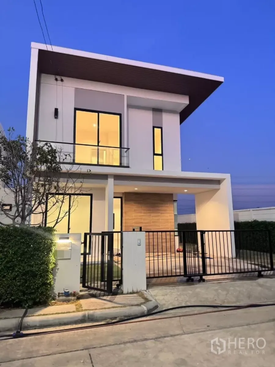Detached House for rent in Samut Prakan - Modern two-storey detached house with slanted roof, warm lights and black gate at dusk.