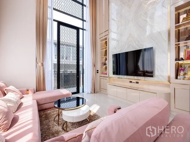 Detached House for sale in Prawet, Bangkok - Double-height living room with marble TV wall, built-in shelving, and pink sectional sofa.