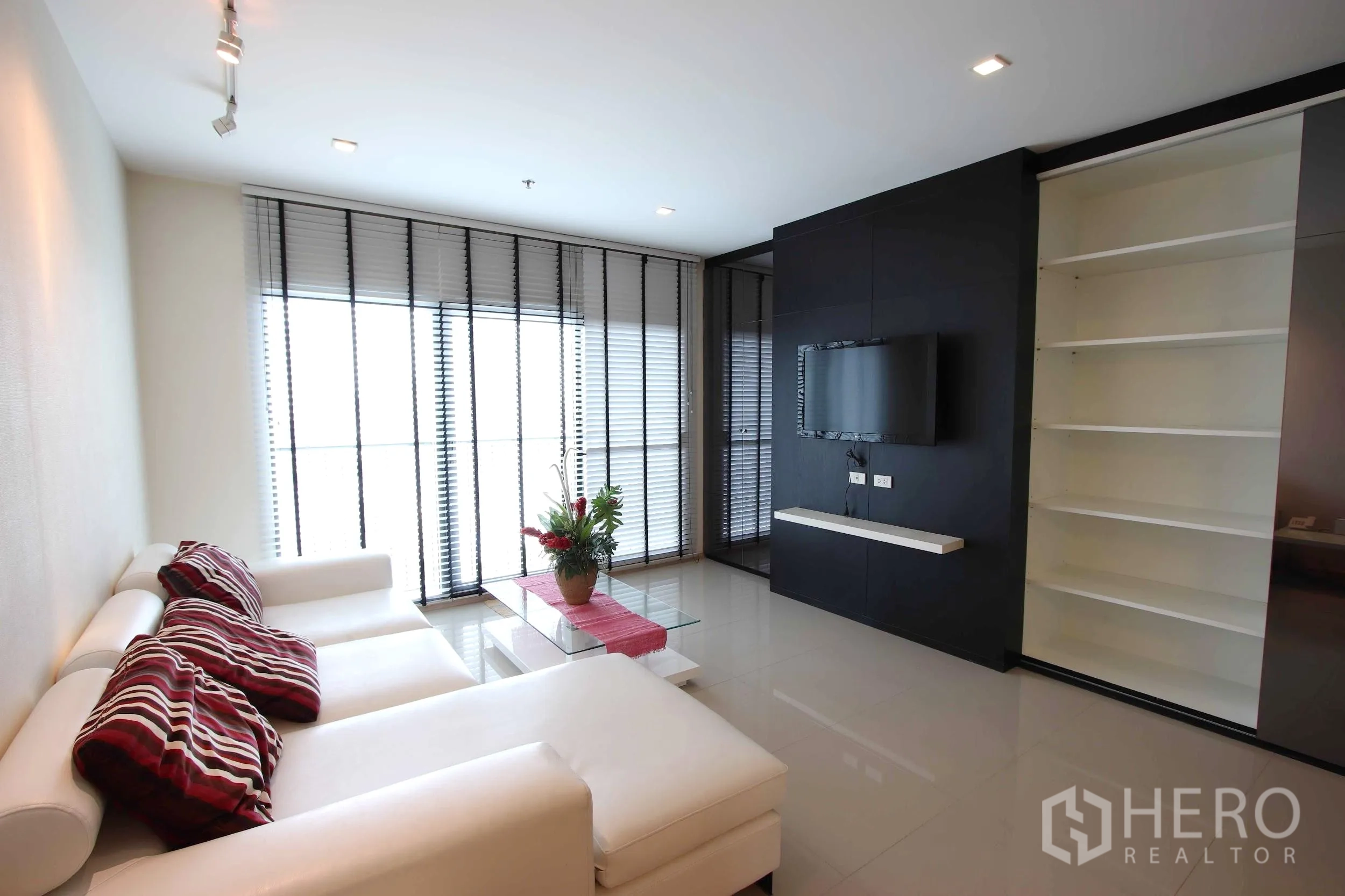 Condominium for rent in Khlong Toei, Bangkok - Bright living room with white sectional sofa, TV wall, built-in shelves and large windows with blinds in a Khlong Toei condominium.