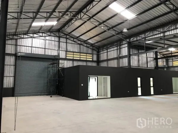 Factory for rent in Bang Yai, Nonthaburi - Interior view showing a roller shutter loading door beside dark grey office rooms under a high ceiling.