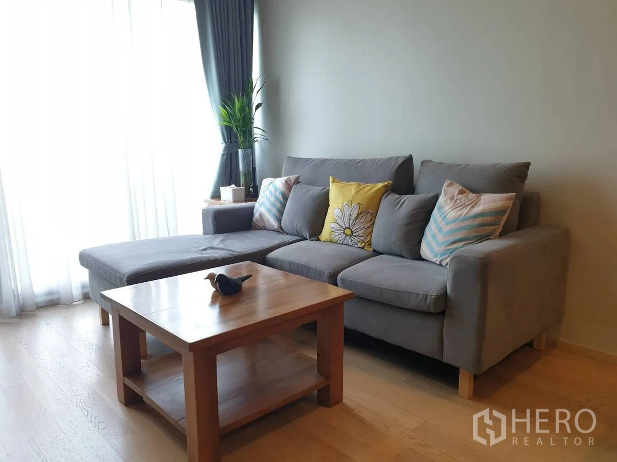 Condominium for rent or sale in Bangkok - L-shaped sofa by large curtained windows with a wooden coffee table and indoor plant.