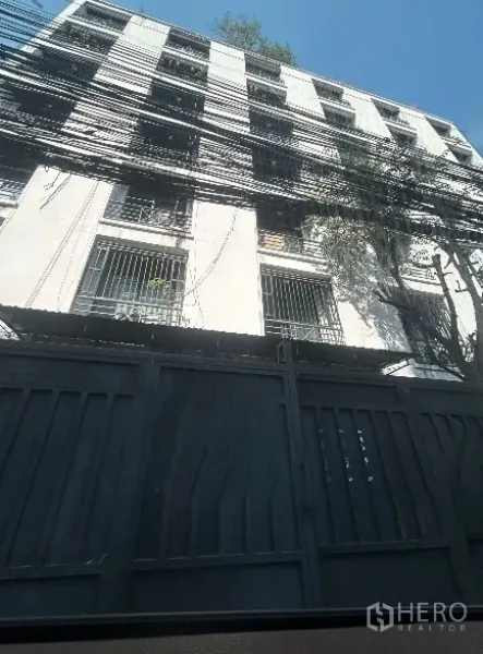 Warehouse for rent in Khlong Toei, Bangkok - Multi‑storey warehouse-style building exterior with barred windows, tall metal gate, and overhead utility cables in urban Khlong Toei under a clear sky.
