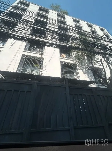 Warehouse for rent in Khlong Toei, Bangkok - Multi‑storey warehouse-style building exterior with barred windows, tall metal gate, and overhead utility cables in urban Khlong Toei under a clear sky.