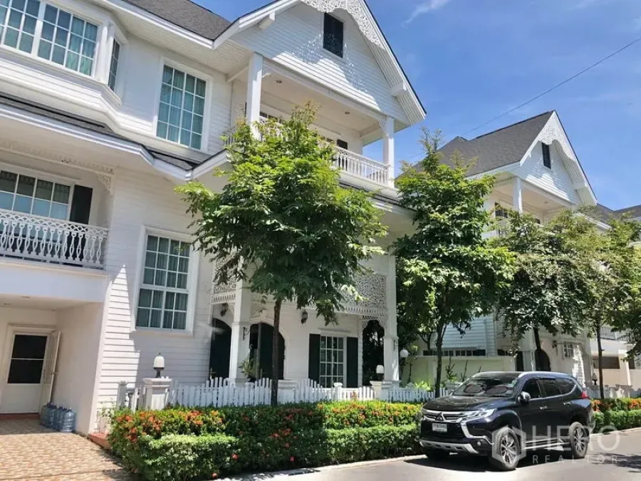 Detached House for rent in Mueang Samut Prakan, Samut Prakan - Multi‑level white colonial‑style detached house with gable roof, balconies, picket fence, trees and driveway with parked SUV.