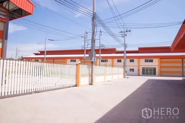 Factory for sale in Mueang Samut Prakan, Samut Prakan - Gated concrete yard of the industrial compound with perimeter fence and matching factory buildings in view.