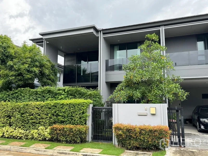 Detached House for rent in Bang Kapi, Bangkok - Modern two‑storey detached house with glass balconies, tall hedge, gated entrance and covered carport.