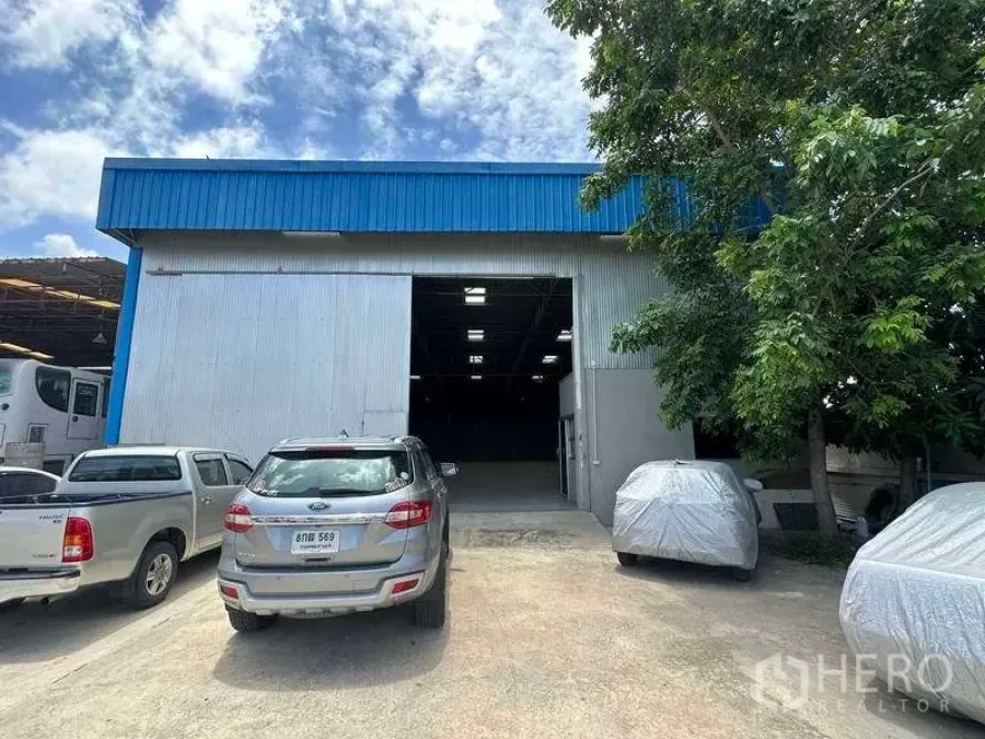货栈 出租在Mueang Pathum Thani, Pathum Thani - 蓝色屋檐金属外墙的仓库外观，卷帘大门开启，前方停车区