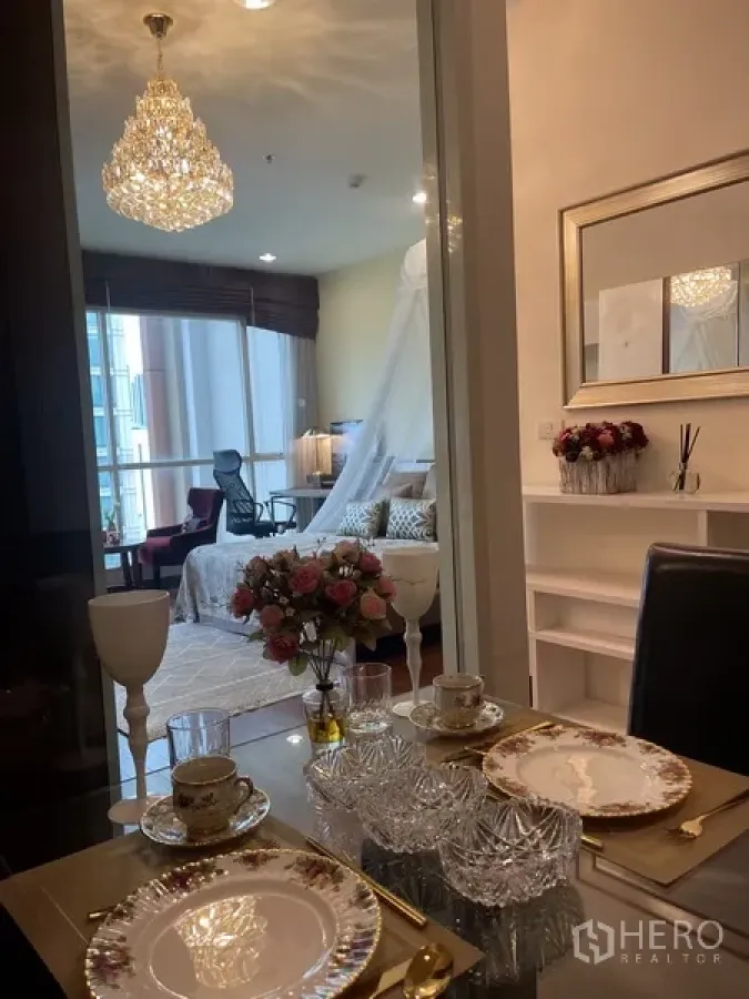 Condominium for rent or sale in Pathum Wan, Bangkok - Elegant dining area with glass table and chandelier overlooking a bright bedroom in a Bangkok condo.