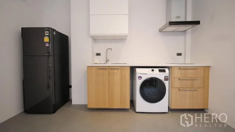 Townhouse for rent in Khlong Toei, Bangkok - Minimalist kitchen with black refrigerator, light wood cabinets, range hood, and washer-dryer.