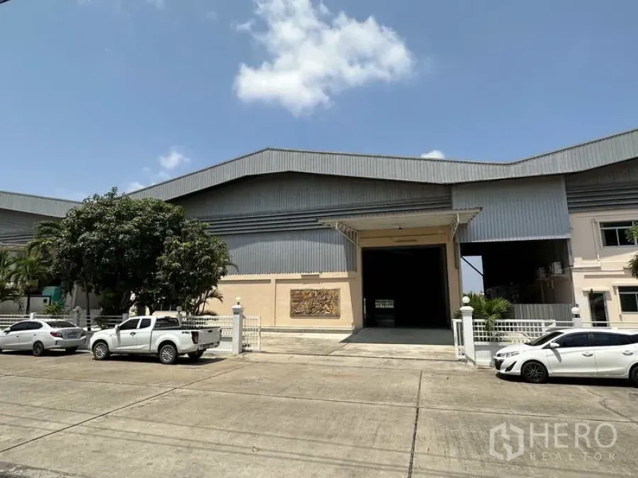 工厂 出租 - 大型金属外墙工厂外观，宽幅卷闸门、围栏入口与停车区，蓝天晴朗。