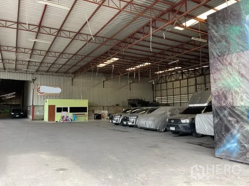 Warehouse for sale in Lam Luk Ka, Pathum Thani - Spacious warehouse interior with high ceiling, small office kiosk, and vehicles parked along the wall.