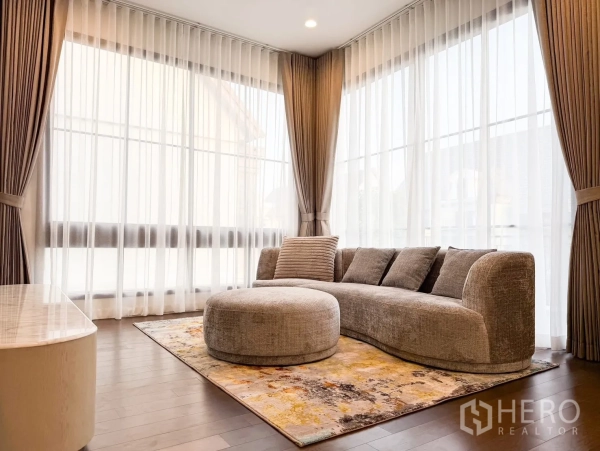 Detached House for rent in Bang Khen, Bangkok - Upstairs lounge with curved sofa, ottoman and wraparound windows with sheer drapes.