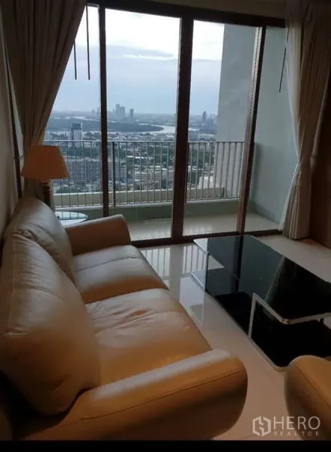 Condominium for rent - Bright living room with beige sofa and glass coffee table opening to a balcony with sweeping Bangkok skyline and river glimpses.