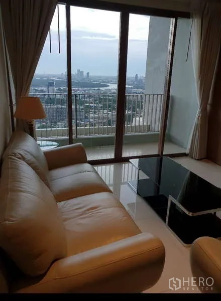 Condominium for rent - Bright living room with beige sofa and glass coffee table opening to a balcony with sweeping Bangkok skyline and river glimpses.