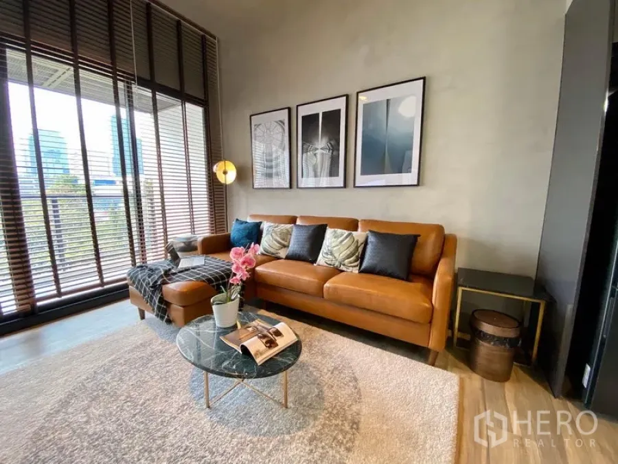 Condominium for rent in Watthana, Bangkok - Bright living room with tan leather sofa, floor-to-ceiling windows with blinds, city view, and round glass coffee table.
