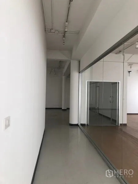办公空间 出租在Bang Rak, Bangkok - 明亮走廊，白色墙面、挑高天花与玻璃隔断，通向木地板办公区。