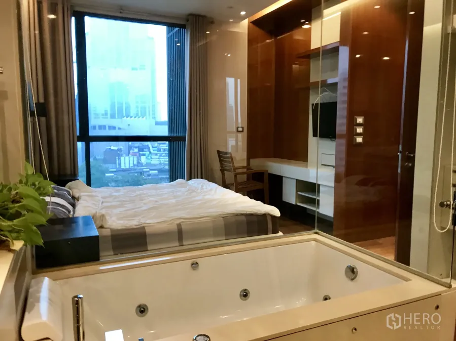 Condominium for rent or sale in Khlong Toei, Bangkok - Bedroom with city view window, built‑in media wall and a glass-partitioned jacuzzi bathtub.