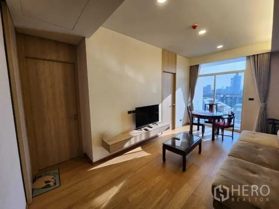 Condominium for rent in Khlong Toei, Bangkok - Open living room with sofa, TV console and round dining set by large window.