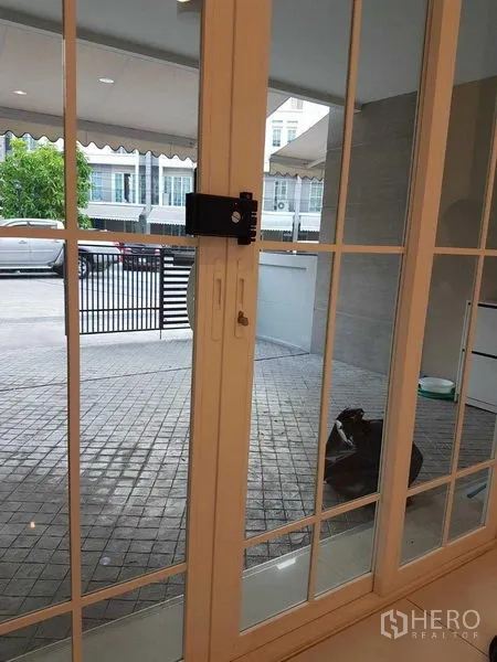 Townhouse for rent in Bang Na, Bangkok - View through white-framed sliding glass door to gated carport and tiled front patio.