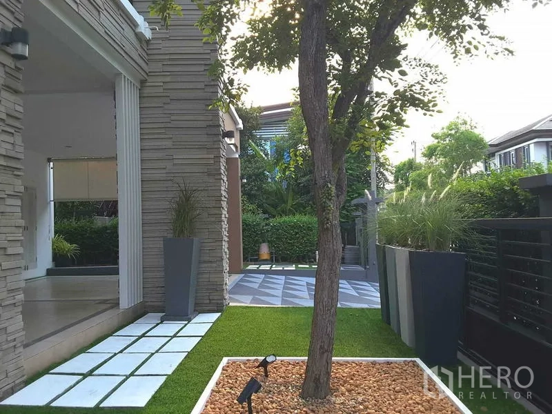 Detached House for rent in Lam Luk Ka, Pathum Thani - Landscaped front garden with a tree, stone-clad pillar and geometric driveway tiles.