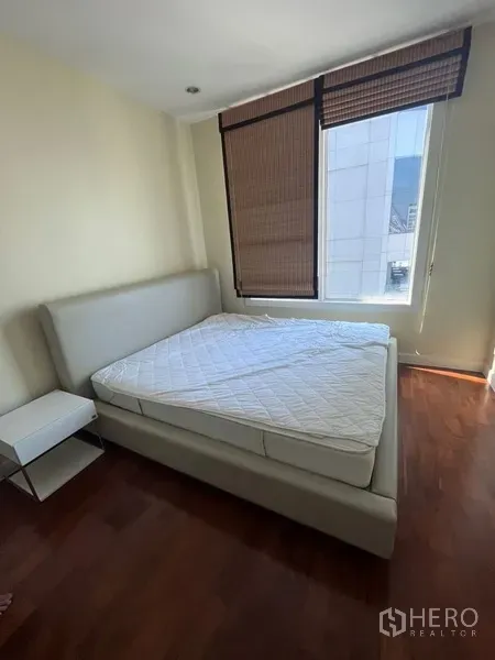 Condominium for rent in Khlong Toei, Bangkok - Cozy bedroom with upholstered bed, bedside table and a large window with blinds over warm wood flooring.
