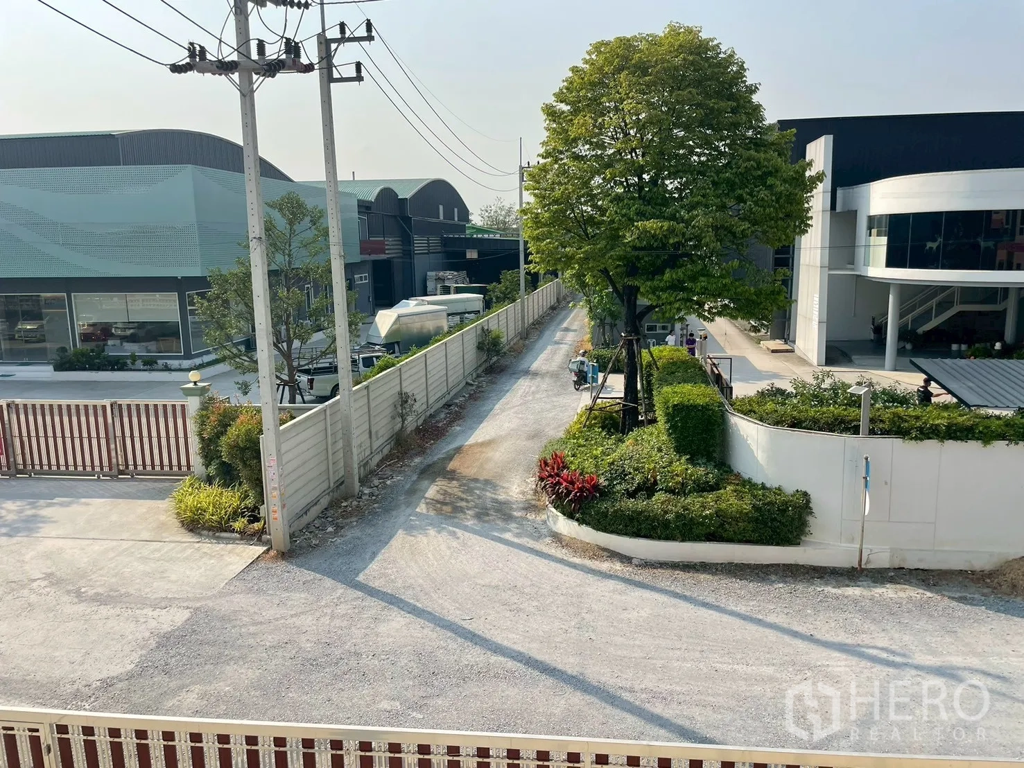 Factory for sale in Sam Phran, Nakhon Pathom - View along the lane with gated industrial buildings, greenery, and utility poles.