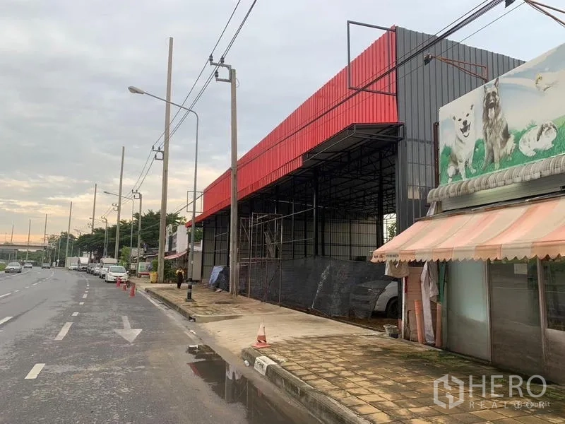 Warehouse for rent in Bueng Kum, Bangkok - Sidewalk view of a high-clearance steel warehouse with red roofline, scaffolding and adjacent shopfronts.