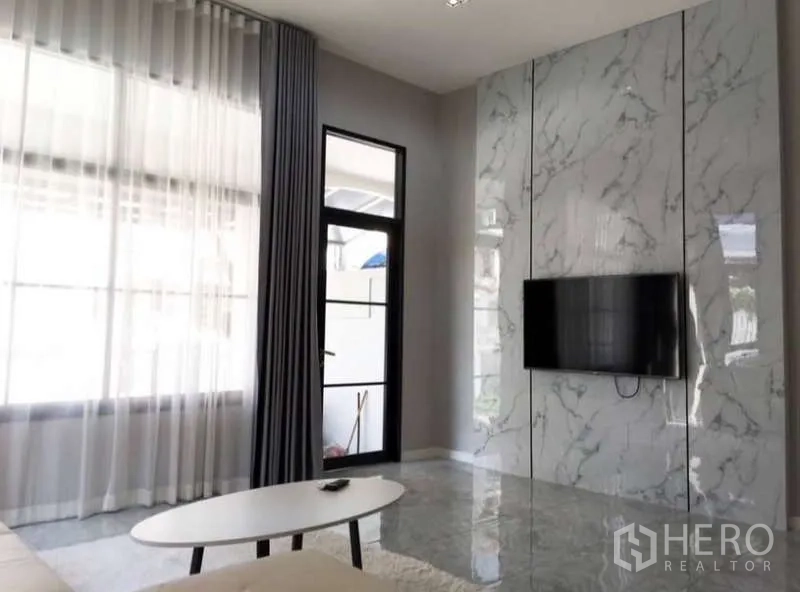 Townhouse for rent or sale in Prawet, Bangkok - Living room TV wall clad in white marble panels with large window and door.