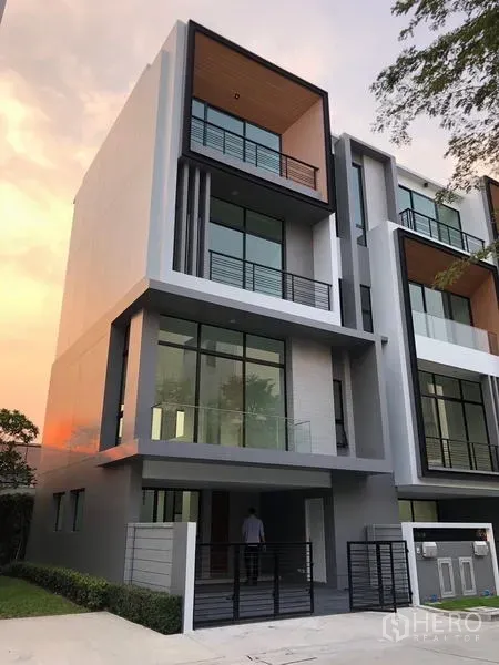 Townhouse for rent or sale in Suan Luang, Bangkok - Modern four-storey townhouse facade with glass balconies at sunset in a gated community.