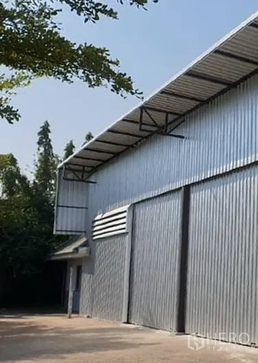 Warehouse for rent in Din Daeng, Bangkok - Side view of the warehouse showing large sliding metal doors and a wide driveway shaded by trees.