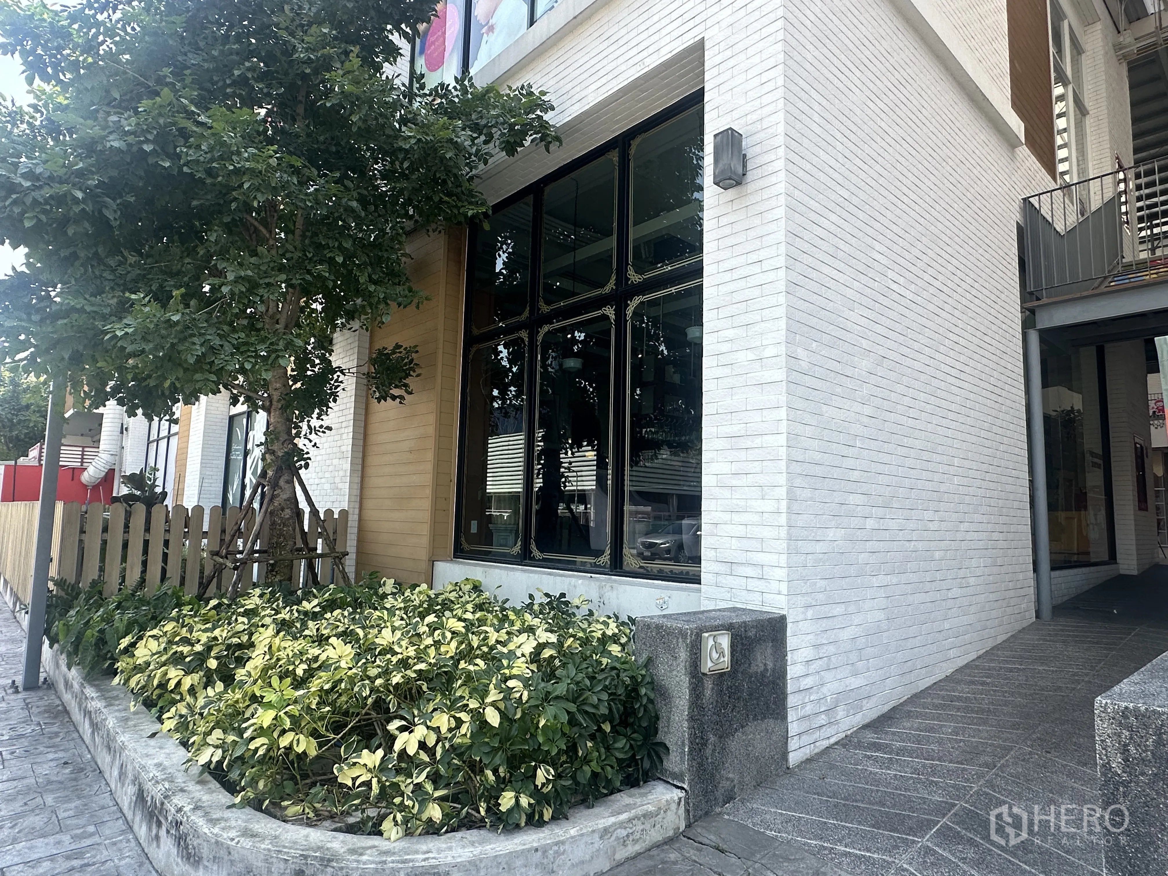 Retail Space for sale in Khlong Toei, Bangkok - Corner of a white brick building with tall windows, landscaped planter with a tree and shrubs, and a side access path.
