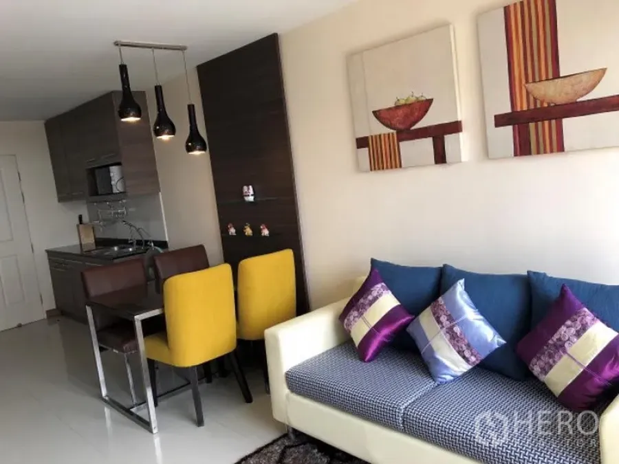 Condominium for rent in Khlong Toei, Bangkok - Open-plan living and dining area with a sofa, colorful cushions, pendant lights, and compact kitchen cabinetry.