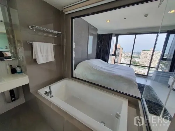 Condominium for sale in Khlong Toei, Bangkok - En-suite bathroom with soaking tub and window to the bedroom, with a city view beyond.
