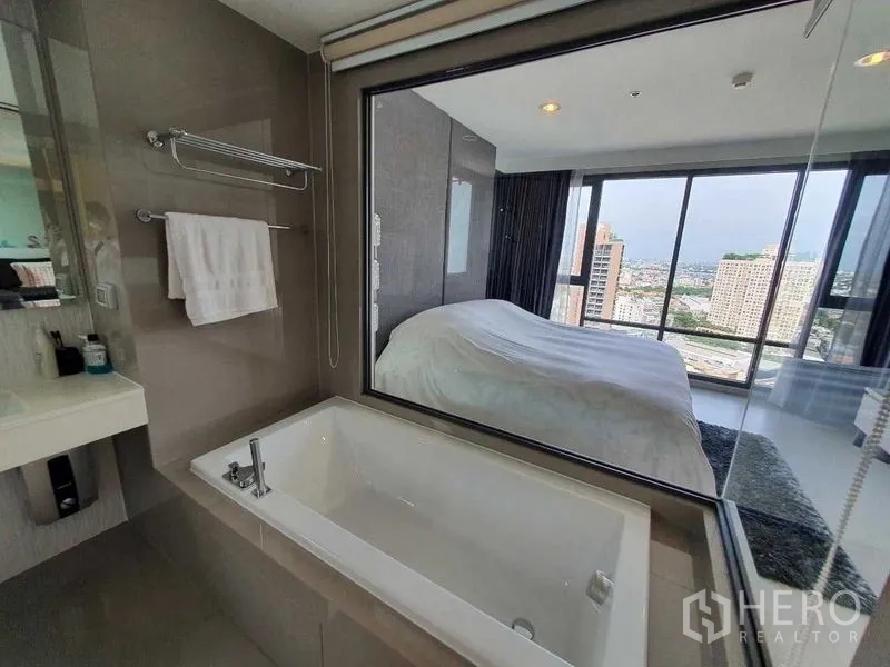 Condominium for sale in Khlong Toei, Bangkok - En-suite bathroom with soaking tub and window to the bedroom, with a city view beyond.