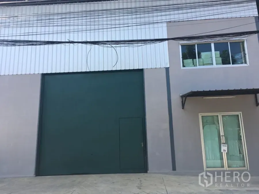 Warehouse for rent in Bang Kapi, Bangkok - Modern warehouse exterior with tall green roller shutter door and a glass-fronted office entry.