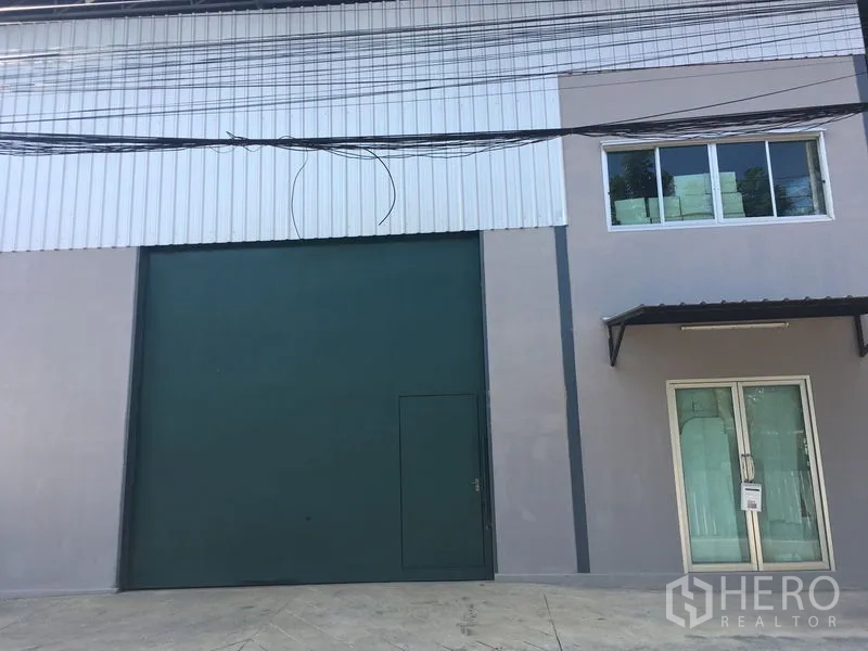 Warehouse for rent in Bang Kapi, Bangkok - Modern warehouse exterior with tall green roller shutter door and a glass-fronted office entry.