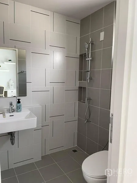 Townhouse for rent in Samut Prakan - Modern bathroom with gray and white geometric tiles, wall-mounted sink, mirror, walk-in shower and toilet.