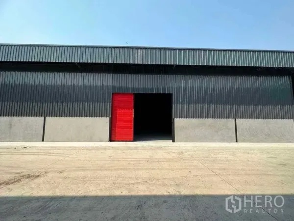 Warehouse for rent in Khlong Sam Wa, Bangkok - Modern black metal warehouse facade with a red roll-up door and open loading bay on a wide concrete yard.