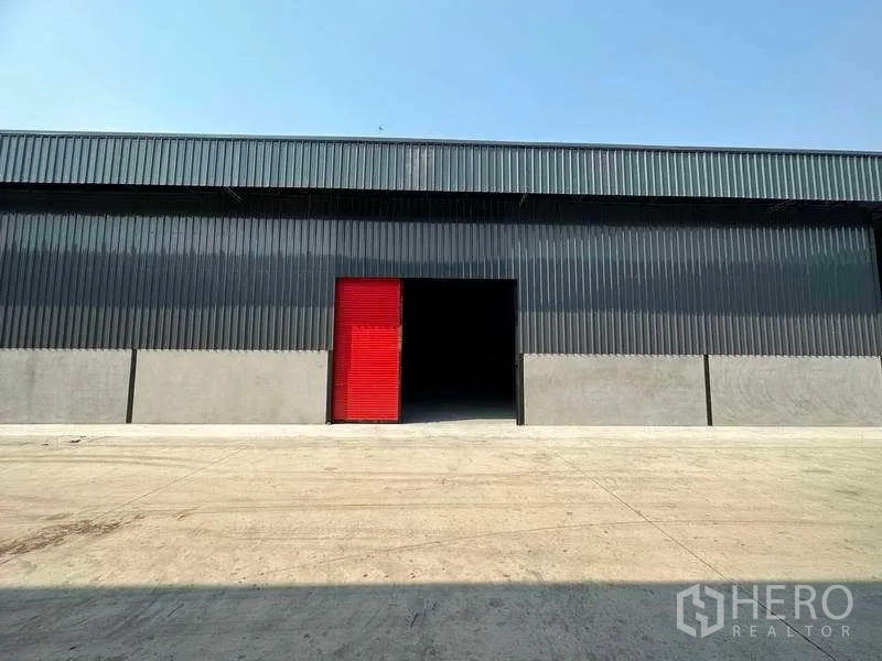 Warehouse for rent in Khlong Sam Wa, Bangkok - Modern black metal warehouse facade with a red roll-up door and open loading bay on a wide concrete yard.