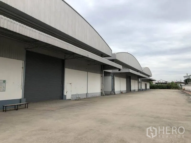 Warehouse for rent in Bang Phli, Samut Prakan - Long warehouse facade with multiple covered loading bays, roll-up doors, and a wide concrete yard for trucks in Bang Phli, Samut Prakan.