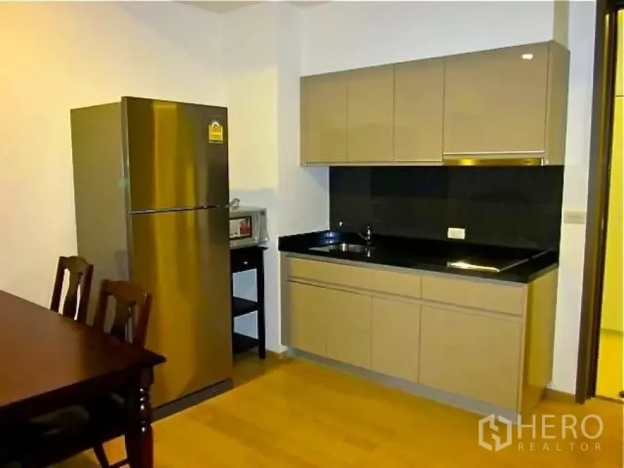 Condominium for rent in Watthana, Bangkok - Compact kitchen with glossy cabinets, black countertop, stainless refrigerator, and dining set.