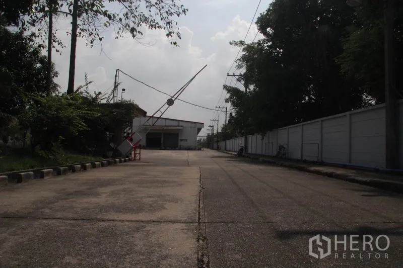 Warehouse for rent in Lam Luk Ka, Pathum Thani - Security gate with barrier arm and private access road into the compound.