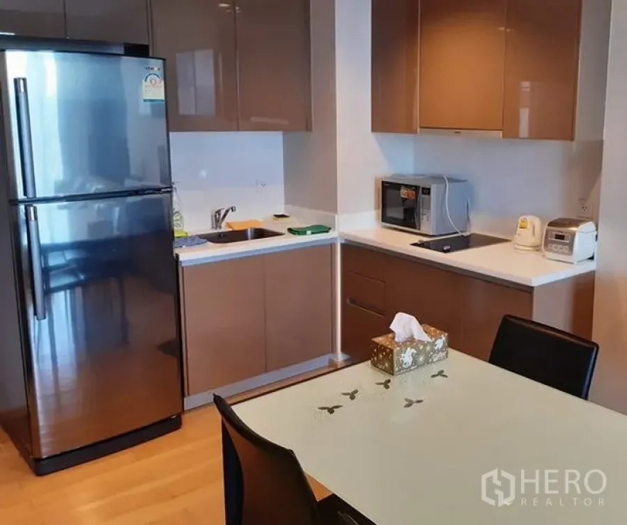 Condominium for rent in Khlong Toei, Bangkok - Modern kitchenette with glossy cabinets, stainless-steel fridge, built-in hob and microwave beside a glass-top dining table.