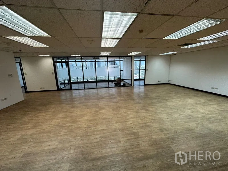 Office Space for rent - Spacious open-plan office with wood floors, grid ceiling lights, and a glass wall facing city views.